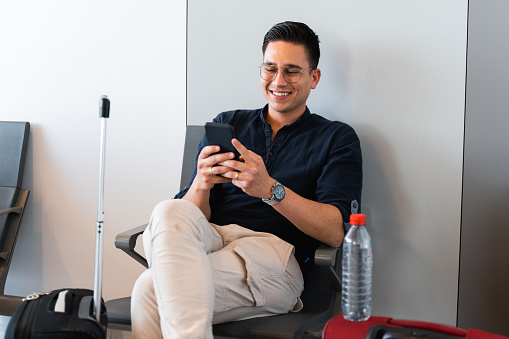 Handsome male with happy face using smartphone while waiting to boarding. Man sitting in lounge airport.