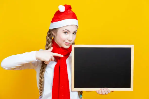 Winsome Caucasian Teenager Girl In Santa Hat and Red Scarf  Holding Black Wooden Blackboard While Pointing With Pointfinger On Yellow Background. Horizontal Image