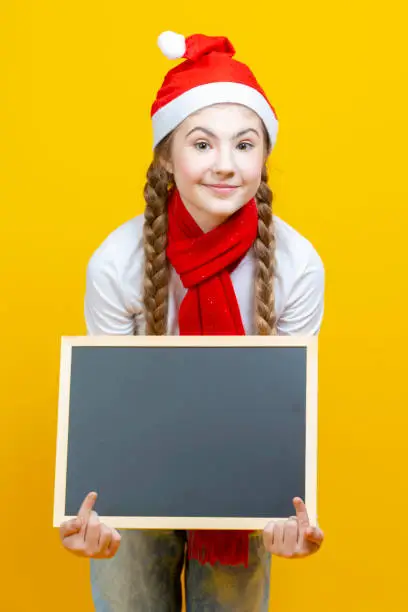 Happy Winsome Caucasian Teenager Girl In Santa Hat and Red Scarf  Holding Black Wooden Blackboard While Pointing With Pointfinger On Yellow Background. Vertical Orientation