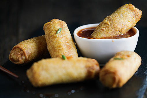 Egg rolls in sweet and sour sauce over a dark rustic table. Garnished with green onions. Selective focus with blurred foreground and background.