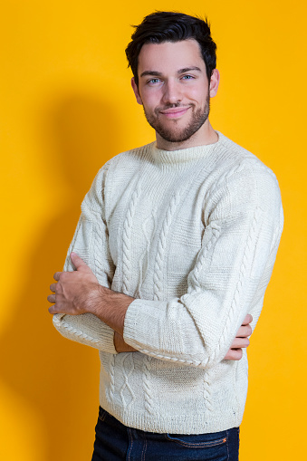 Smiling Caucasian Handsome Man In knitted White Striped Sweater Posing Against Yellow Background While Smiling Happily With Hands Folded. Vertical Image