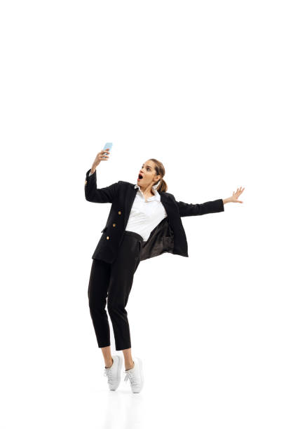 若い女性のダイナミックな肖像画、黒い公式スーツを着たオフィスワーカーが、白いスタジオの背景に隔離された電話でビデオ通話をしている - jumping business women dancing ストックフォトと画像
