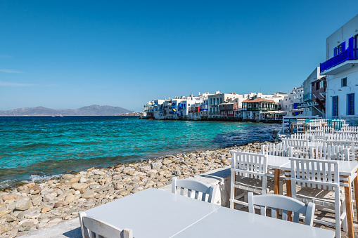 Empty tables and chairs of Greek restaurants and cafes at the waterfront of Little Venice, a romantic and famous travel location in Mykonos, Mykonos Island, Greece. Colorful image with blue sky in summer.