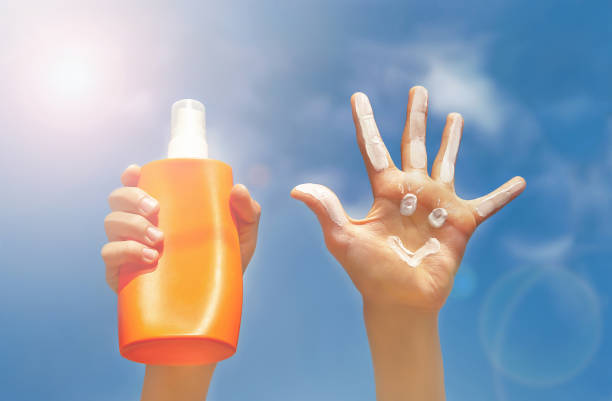 Two Caucasian hands on blue sky, clouds and beams of sun background. Positive symbol drawing by sunscreen (sun cream) on open hand, another hand holding bottle of suntan lotion. Concept of protection from sun, skin care.