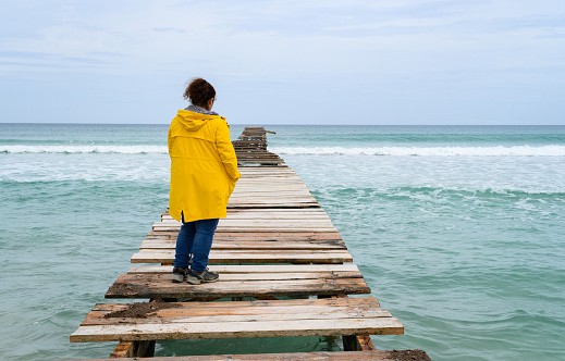 woman standing on old wooden pier with yellow raincoat looking out to sea. Concept of traveling in freedom