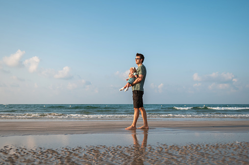 Man holding his baby boy by the seaside on the beach on a summer vacation. Father and son on the seaside spending time outdoors