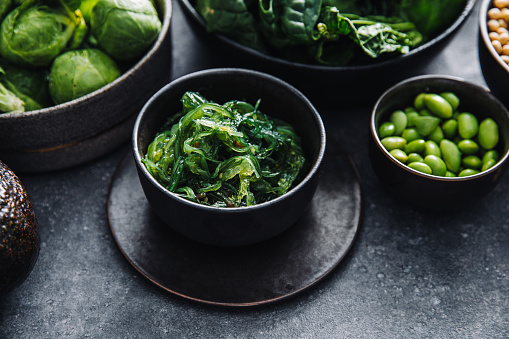 Close-up of healthy raw food in bowls on black table. Fresh green spinach, edamame, wakame, and brussel sprouts in bowls on a table.