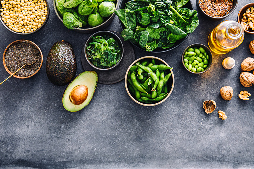 Variety of Omega 3 vegan food items on black table. Table top view of fresh spinach, flax seeds, walnuts, walnut oil bottle,  brussel sprouts, lentil seeds, chia seeds, avocado, edamame and wakame in bowls on a table.