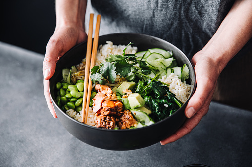 Close-up of a woman holding a poke salad bowl with chopsticks. Female hands with healthy salad over table.