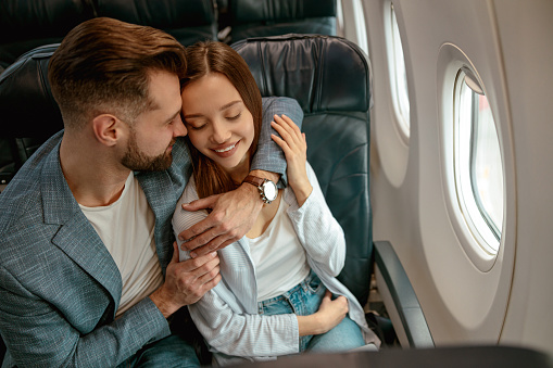Joyful woman sitting in passenger chair and smiling while loving man embracing her during flight
