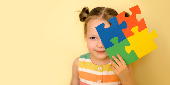 A cute beautiful blue - eyed girl holds an autism symbol in hand, covering her face with it and smiling sweetly. Conceptual photo for Autism Day