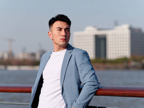 Portrait of handsome Chinese young man in light blue suit and white undershirt looking away with modern city buildings background in sunny day, front view of confident businessman. stock photo
