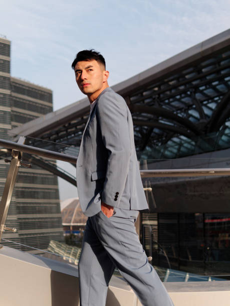Portrait of handsome Chinese young man in light blue suit and white undershirt walking and looking away with modern city buildings background in sunny day, side view of confident businessman. stock photo