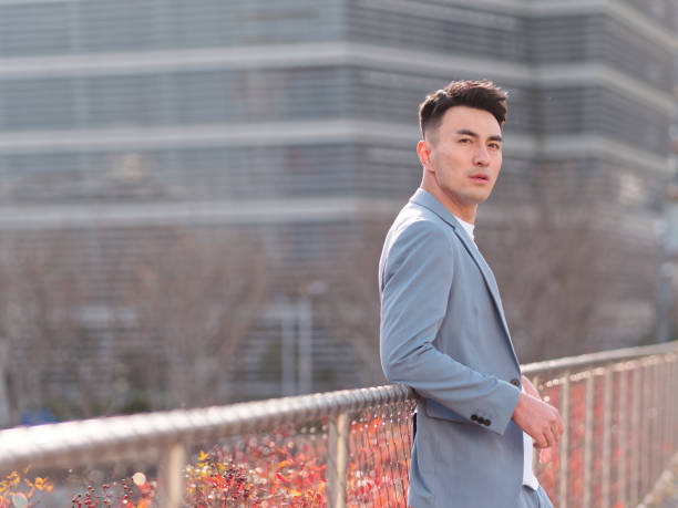 Portrait of handsome Chinese young man in light blue suit looking at camera with city buildings background, side view of confident businessman. stock photo