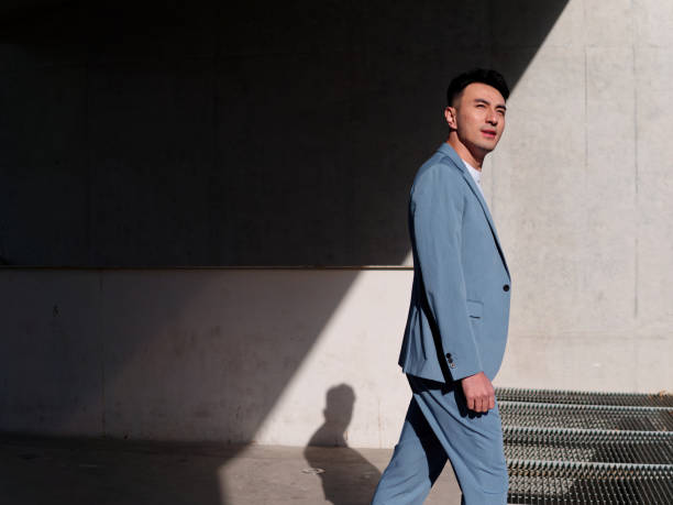 Confident businessman, portrait of handsome Chinese young man in light blue suit smiling and walking into sunlight, side view. stock photo