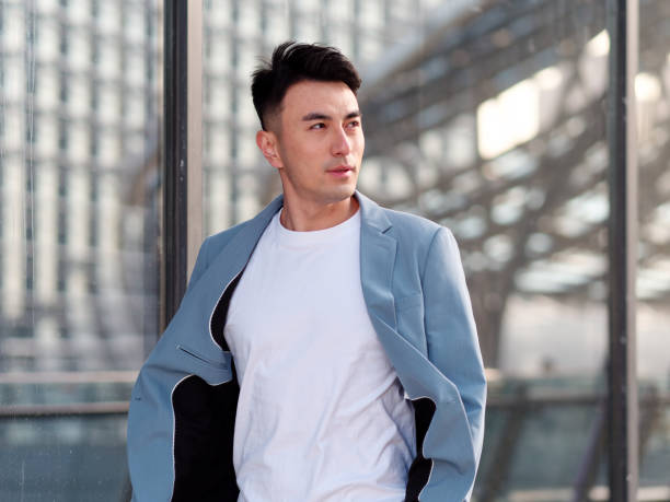 Portrait of handsome Chinese young man in light blue suit and white undershirt walking and looking away with modern city buildings background in sunny day, front view of confident businessman. stock photo
