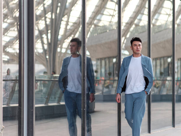 Portrait of handsome Chinese young man in light blue suit and white undershirt walking and looking away with his reflections in glass wall background in sunny day, front view of confident businessman. stock photo