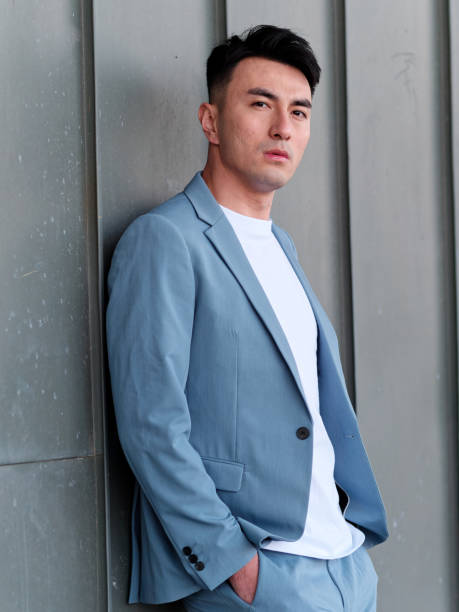 Portrait of handsome Chinese young man in light blue suit and white undershirt leaning on wall with hands in pockets and looking at camera with serious expression, front view of businessman. stock photo