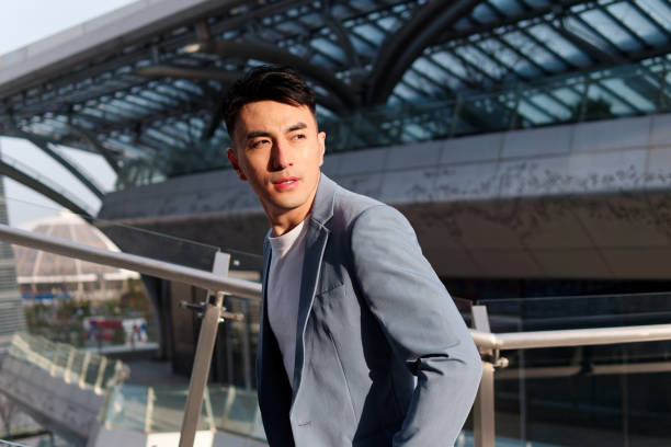 Portrait of handsome Chinese young man in light blue suit and white undershirt walking and looking away with modern city buildings background in sunny day, side view of confident businessman. stock photo