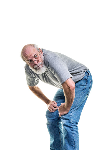 A casually dressed, frustrated? angry? confused? defeated? senior adult man is bending over - leaning forward at the waist with his hands on his knees - and staring directly at the camera.