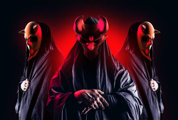 Occult horror sectarians with metal masks on black background. Scary horror occult sectarians in black hood and metal masks on black background with red glow. ritual mask stock pictures, royalty-free photos & images