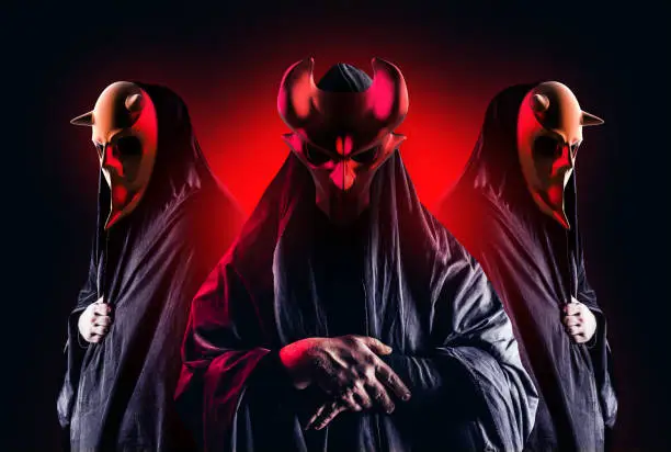 Scary horror occult sectarians in black hood and metal masks on black background with red glow.