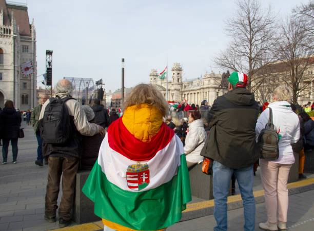 Caucasian White People Celebrating and Commemorating 19th Century National History with Hungarian Flags on the National Day in Kossuth Lajos Square, People Listening to Solemn Historical Memorial Show in the Foreground, Prime Minister Viktor Orbán Also Giving Solemn Speech on Stage in the Background, Hungarian Parliament Building Neighborhood (Left), in 5th District of Downtown Budapest, Hungary, Europe in March 2022