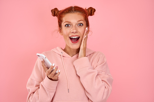 Smiling cheerful red-haired caucasian girl cried out joyfully holding a modern smartphone, recieves unexpected message or news, has overjoyed expression. Concept of great offers and special promotions