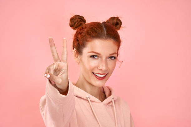 istock Glad ginger woman makes peace victory gesture or two with isolated on pink background 1387753994