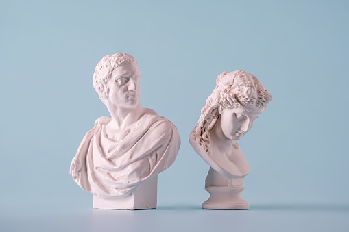 Two small white antique style Roman or Grecian busts depicting men over a blue background