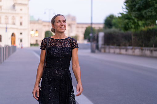 medium shot happy smiling mature adult woman in her forties in beautiful black cocktail dress walking alone through old town of european city going out, shallow focus, copy space