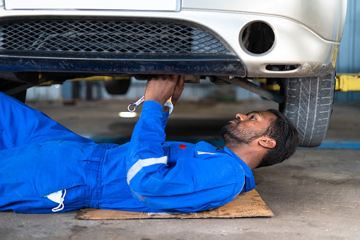 Professional car mechanic lying down while working under car at garage - concept of hard worker, skill labor, blue collar jobs and repair service.