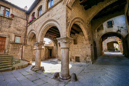 An idyllic view of the ancient stone portico in the square of San Pellegrino, in the medieval heart of Viterbo, in central Italy. The medieval center of Viterbo, the largest in Europe with countless historic buildings, churches and villages, stands on the route of the ancient Via Francigena (French Route) which in medieval times connected the regions of France to Rome, up to the commercial ports of Puglia, in southern Italy, to reach the Holy Land through the Mediterranean. Located about 100 kilometers north of Rome along the current route of the Via Cassia, Viterbo is also known as the city of the Popes, because in the 13th century it was the Papal See for 24 years. Founded in the Etruscan era and recognized as a city with a papal document from the year 852 AD, the entire city is characterized by its stone and tuff constructions, materials abundantly present in this region of central Italy and which have always been used for the construction of houses and churches. Super wide angle image in high definition format.
