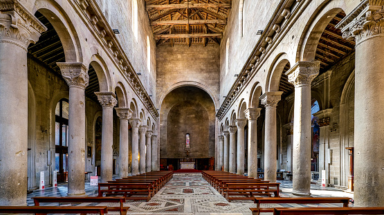 The majestic and imposing central nave and apse of the Duomo of San Lorenzo (Cathedral of Saint Lawrence), in the heart of the medieval city of Viterbo, in central Italy. Built in the Romanesque style in the 12th century, this cathedral became important in the history of the Catholicism when in the 13th century Viterbo became the seat of the Roman Pontiffs. The medieval center of Viterbo, the largest in Europe with countless historic buildings, churches and villages, stands on the route of the ancient Via Francigena (French Route) which in medieval times connected the regions of France to Rome, up to the commercial ports of Puglia, in southern Italy, to reach the Holy Land through the Mediterranean. Located 100 kilometers north of Rome along the current route of the Via Cassia and recognized as a city with a papal document from the year 852 AD, Viterbo is characterized by its stone and tuff constructions, materials abundantly present in this region of central Italy and which have always been used for the construction of houses and churches. Super wide angle image in high 16:9 and definition format.