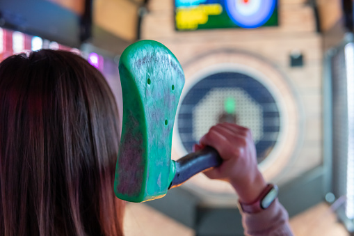 Woman's hand from behind holding a green rubber axe to throw it at the target with the intention of sticking it in the bullseye.