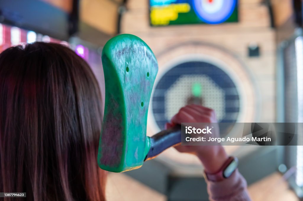 Rear view of a woman holding an axe for recreational throwing in an arcade. Woman's hand from behind holding a green rubber axe to throw it at the target with the intention of sticking it in the bullseye. Axe Stock Photo