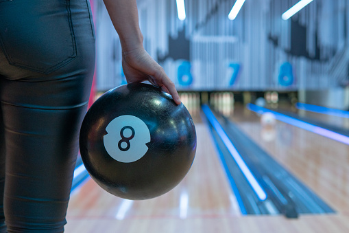 Image of a right hand holding a black bowling ball near the bowling alley that is out of focus.