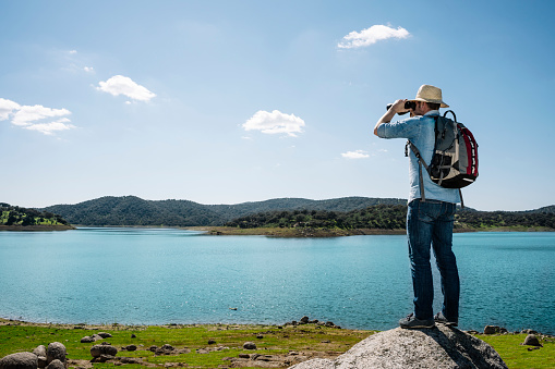 TOURIST MAN ON TOP OF A ROCK WITH BACKPACK AND HAT LOOKING WITH BINOCULARS IN A BEAUTIFUL LANDSCAPE WITH MOUNTAINS NEXT TO A LAKE, RIVER OR SEA. TOURISTIC VIEWPOINT. TRAVELER MAN CONCEPT.