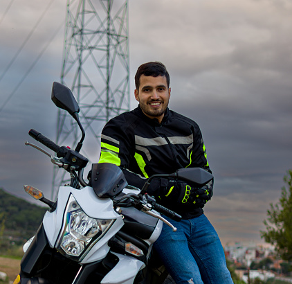 A 3/4 length image of a smiling latin adventure moto-rider leaning on his motorcycle at the sunset in a lookout point of a city