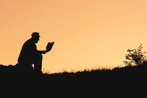 Photo of silhouette of man sitting on rock and reading book. reading books in nature