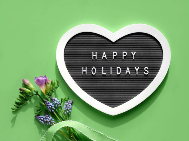 istock Happy Holidays text on felt heart shaped letter board, letterboard. Spring flat lay on light green. Pink flowers 1387822574