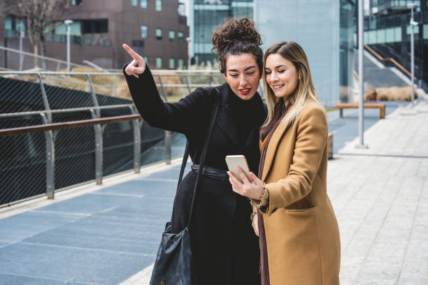 dos mujeres jóvenes que viajan por la ciudad, usando un teléfono inteligente para buscar información y mapas, señalando el lugar que están buscando - italiano idioma fotografías e imágenes de stock