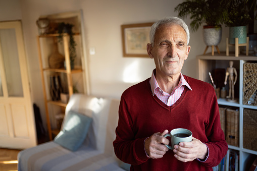 Portrait of pensive Caucasian senior man, looking through the window while holding cup of coffee