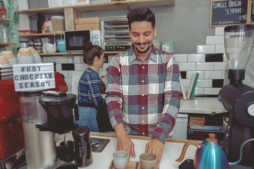Portrait of happy man barista in plaid shirt standing behind bar counter with wooden tray and two cups of coffee on it