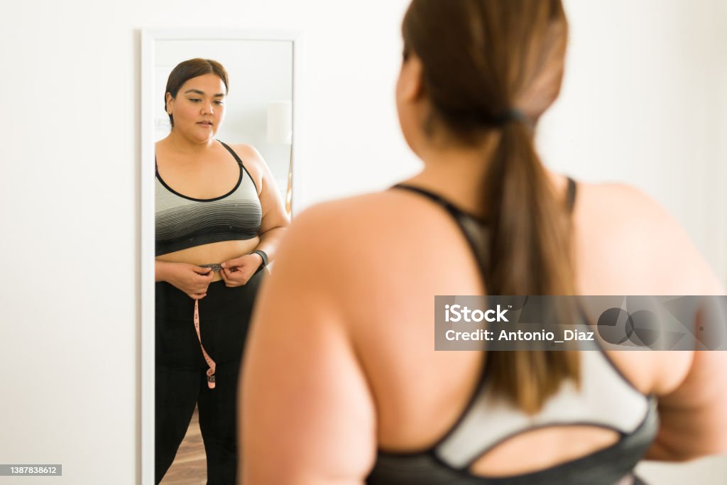 Obese woman with a body positivity concept Overweight woman in her 20s measuring her body and looking in the mirror. Fat latin woman trying to lose weight with exercise and diet Mirror - Object Stock Photo