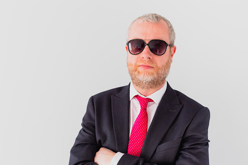 Close up color image depicting a cool mid adult businessman wearing a suit jacket, white shirt and pink necktie and aviator-style sunglasses. White wall background with room for copy space.