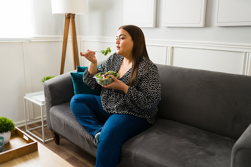 Curvy young woman sitting on the sofa and eating a green salad. Fat woman following a healthy diet to lose weight
