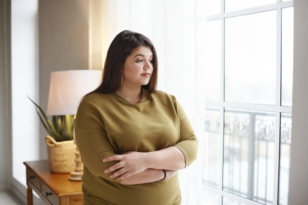 people, lifestyle and body positivity concept. beautiful young brunette female with extra pounds standing by window at home, crossing arms on her chest, having thoughtful pensive look, pondering - overweight imagens e fotografias de stock