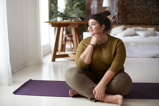 Healthy active lifestyle, fitness, pilates, dieting and excess weight concept. Charming barefooted plus size young woman in sportswear sitting cross legged on mat, going to practice yoga, smiling