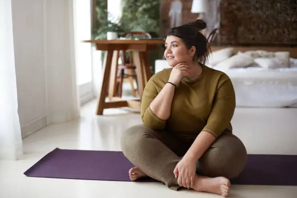 Photo of Healthy active lifestyle, fitness, pilates, dieting and excess weight concept. Charming barefooted plus size young woman in sportswear sitting cross legged on mat, going to practice yoga, smiling